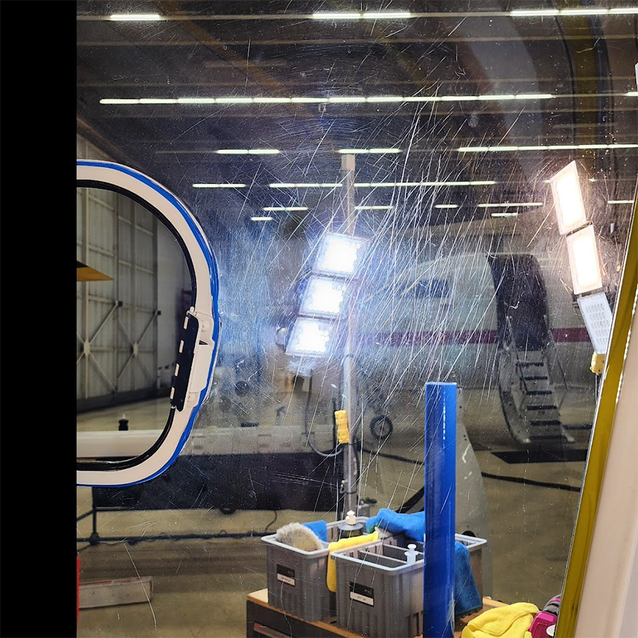 Plane being cleaned inside a hangar. Water streaks across a window, revealing an aircraft and equipment.