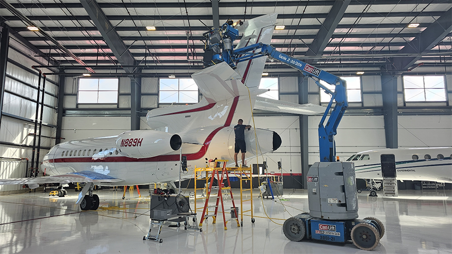 Workers servicing a white jet in a hangar, using a lift. Blue lift, metal scaffolding, and windows are visible.