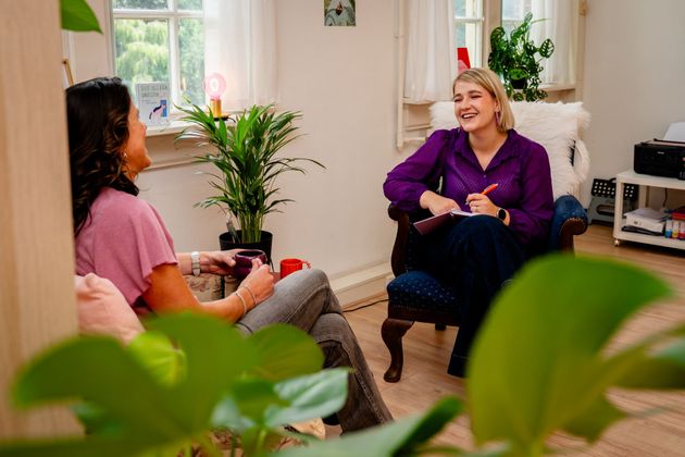 Two women talking in a bright living room, one taking notes in a chair near a window and plants.