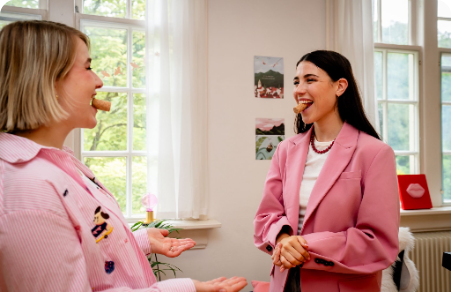 Two women in pink jackets smiling and talking in a bright office with large windows