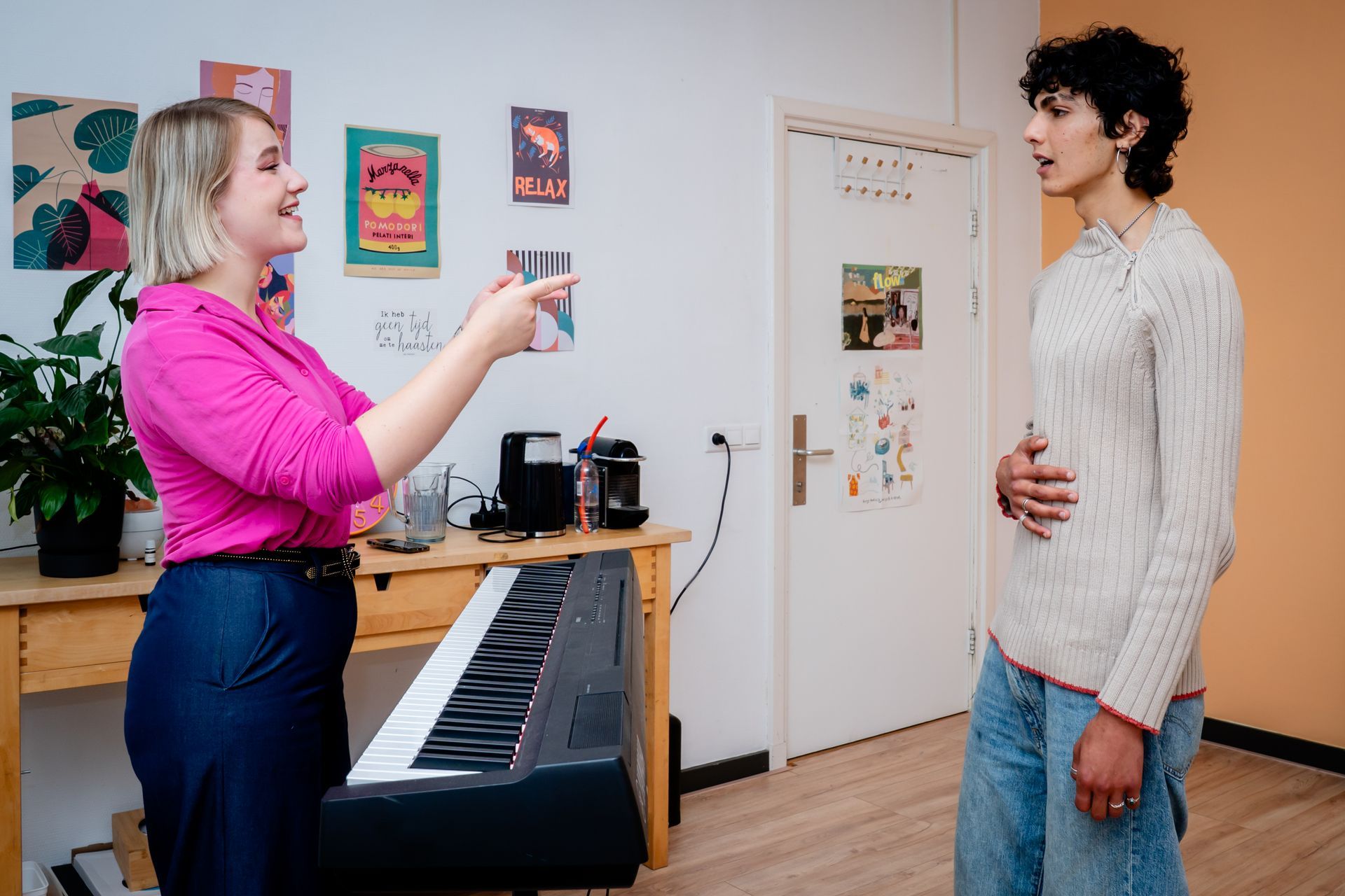 Two women talking in a bright living room, one taking notes in a chair near a window and plants.