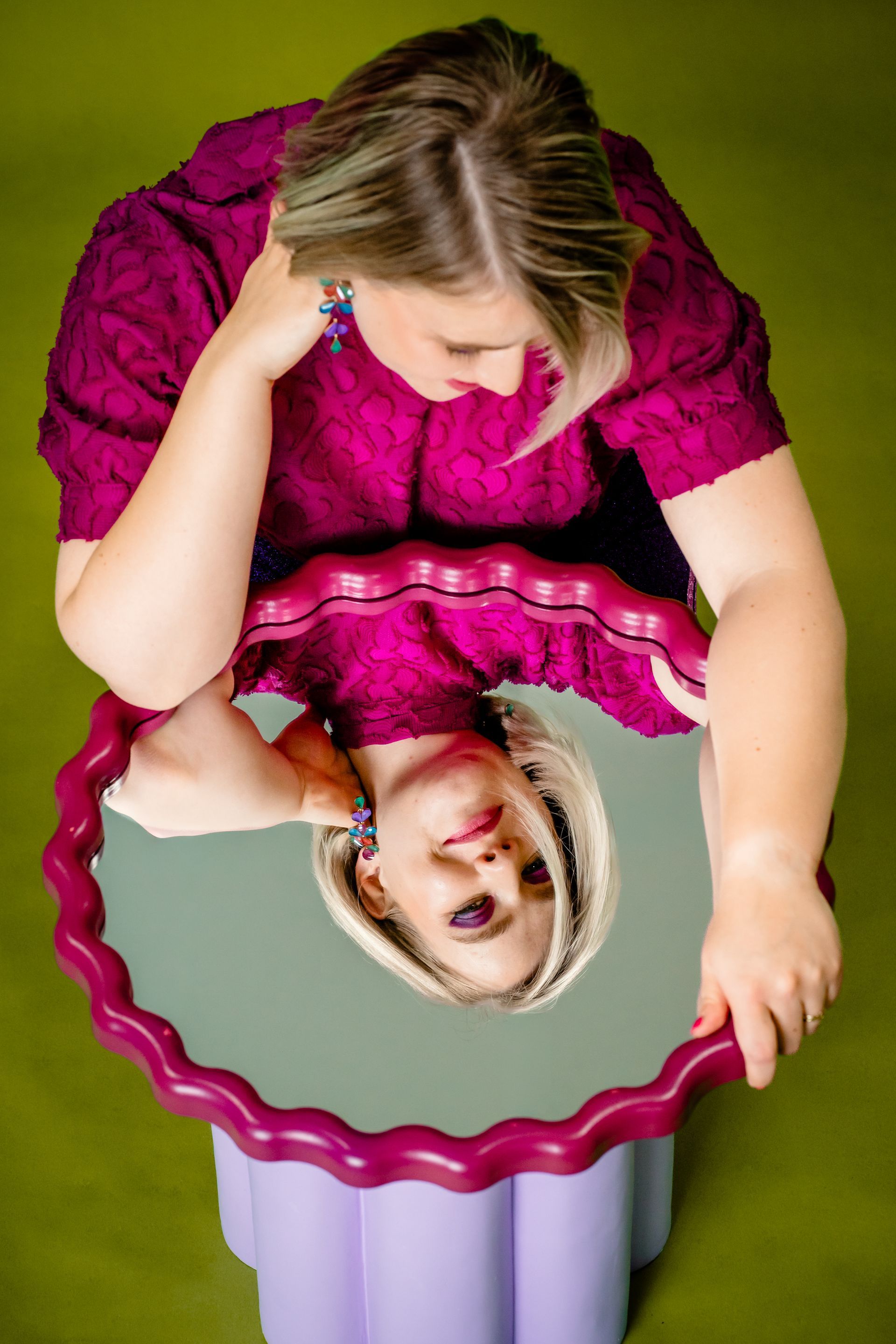 Woman holding a pink wavy mirror, reflecting her smiling image on a magenta background.