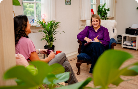 Two women talking in a bright living room, one taking notes in a chair near a window and plants.