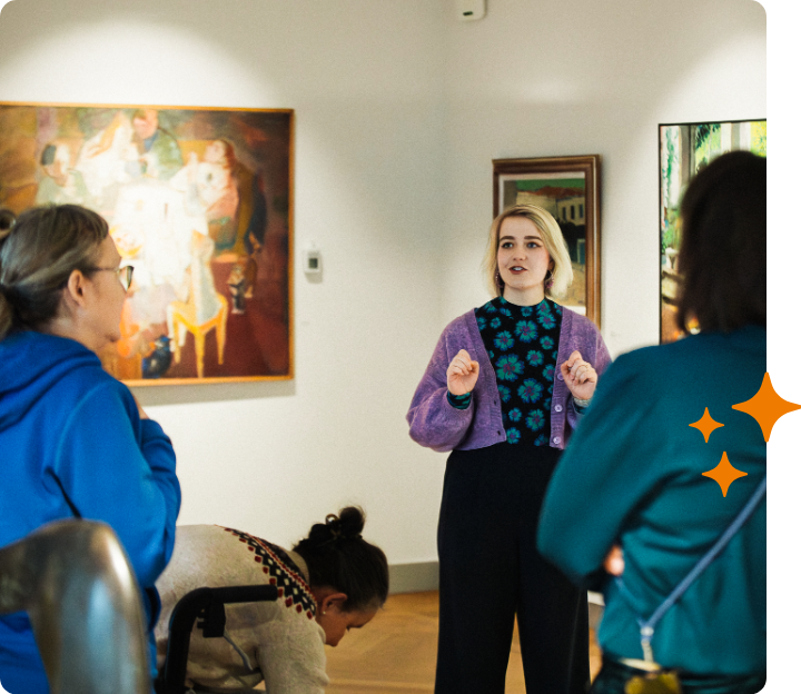 Tour guide speaking in an art gallery to a small group of visitors, with paintings on the walls.