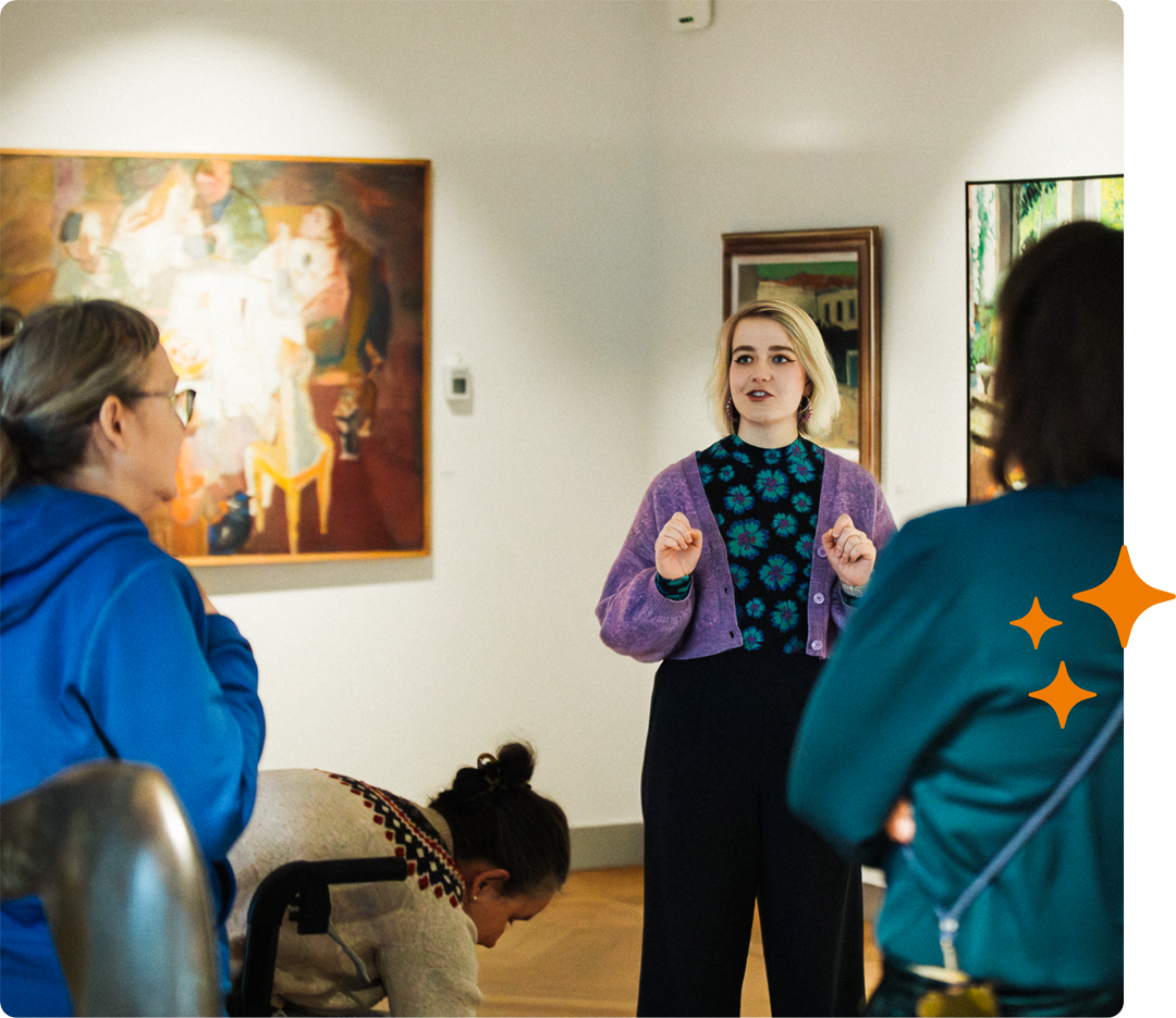 Tour guide speaking in an art gallery to a small group of visitors, with paintings on the walls.