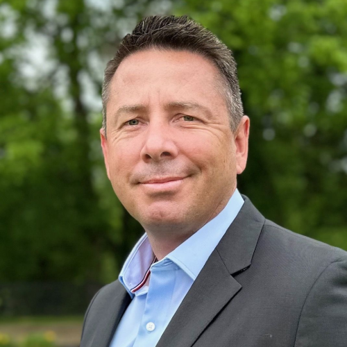 Portrait of a man in a gray suit and light blue shirt outdoors, with green trees blurred behind him