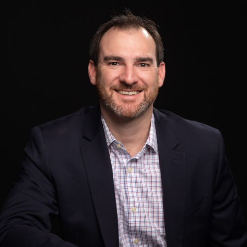 Portrait of a smiling man in a dark blazer and checkered shirt against a black background