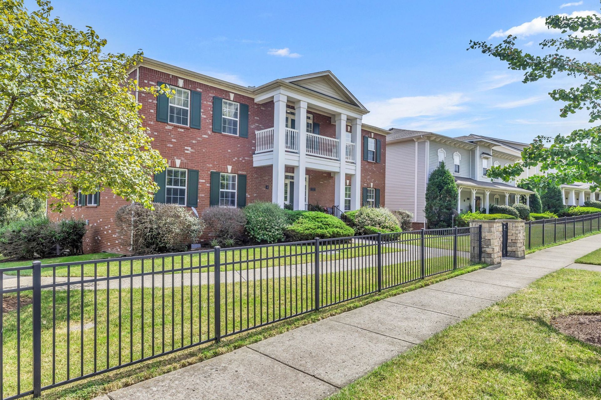 Brick apartment building with white columns, black fence, and sidewalk on a sunny day