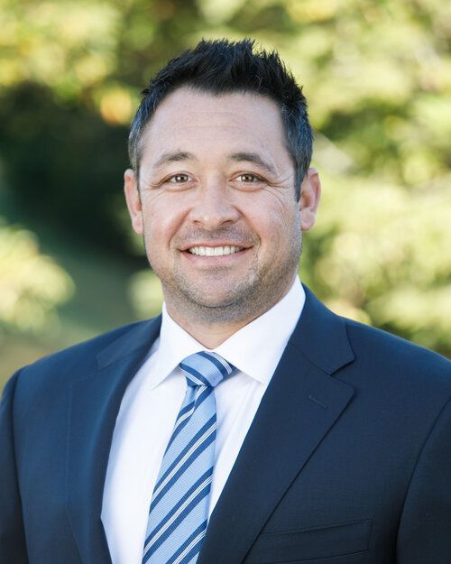 A professional portrait of a smiling person wearing a dark blue suit, white shirt, and blue-striped tie outdoors.