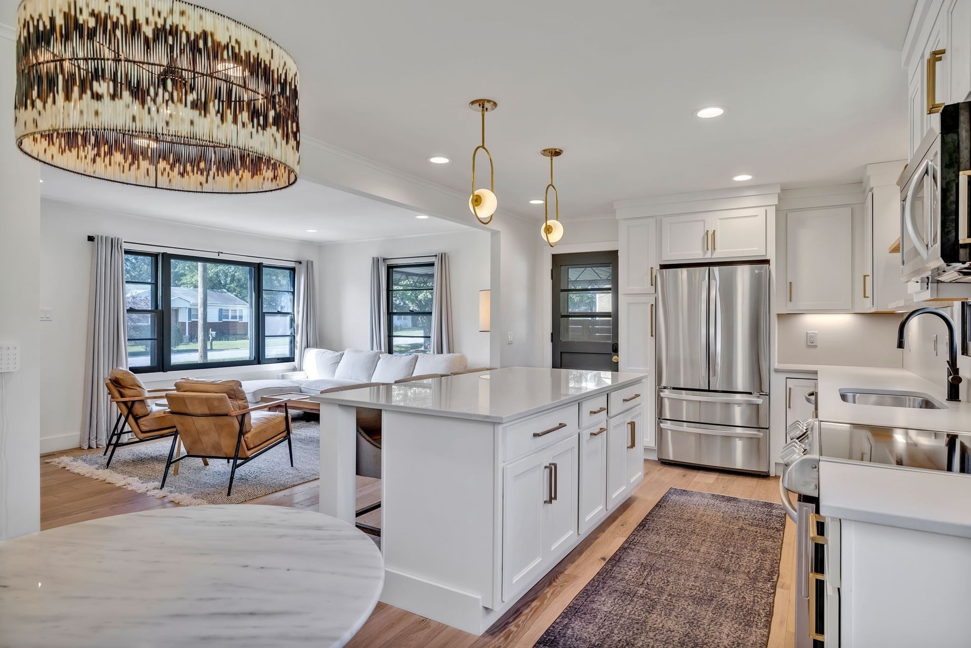 Bright modern kitchen with white cabinets, island, and stainless steel refrigerator, opening to a cozy living room