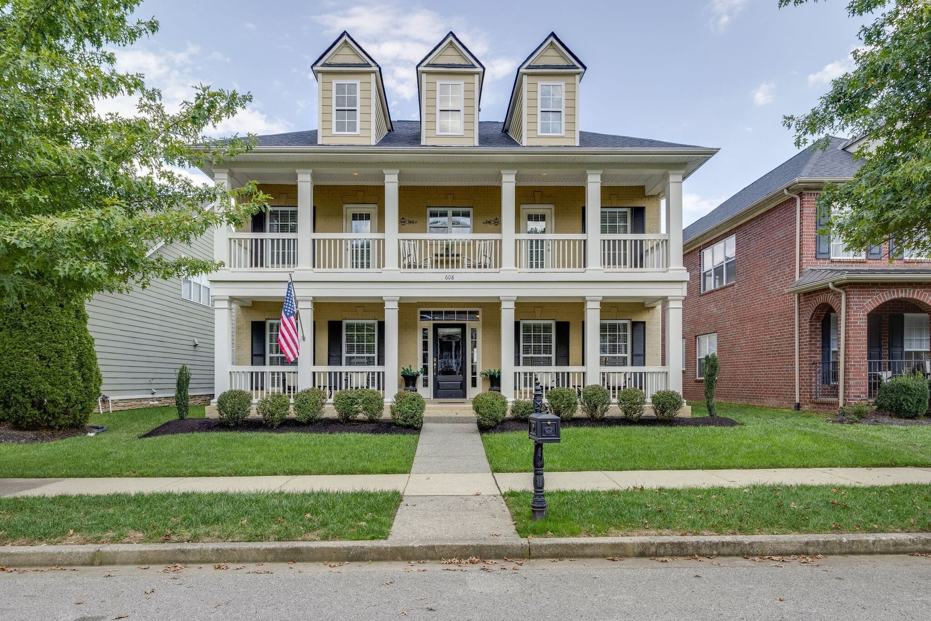 A two-story, yellow colonial-style house with a front porch, balcony, and three dormers, situated on a green lawn.