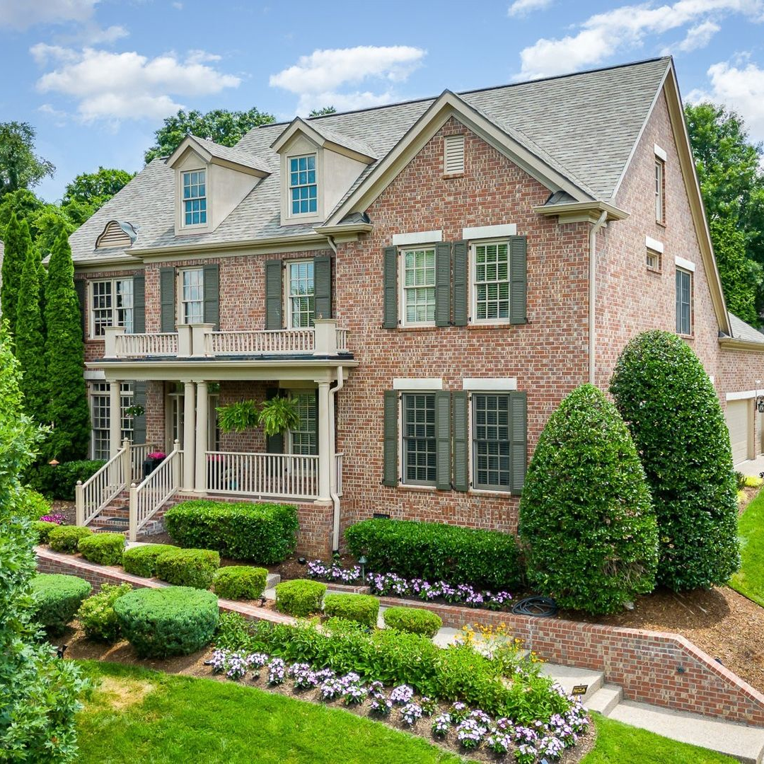 A two-story brick house with a front porch, grey shingled roof, two dormer windows, and manicured landscaping.