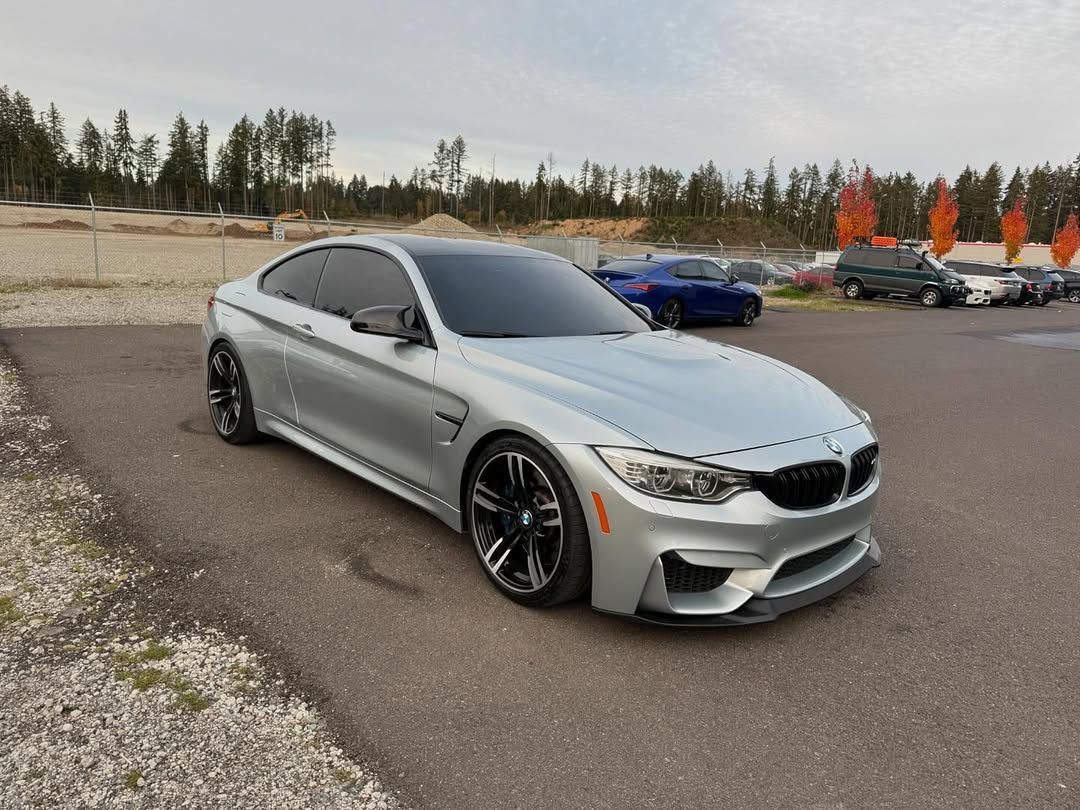 Silver BMW M4 coupe parked on asphalt. Black wheels, tinted windows. Cloudy sky, trees in background.