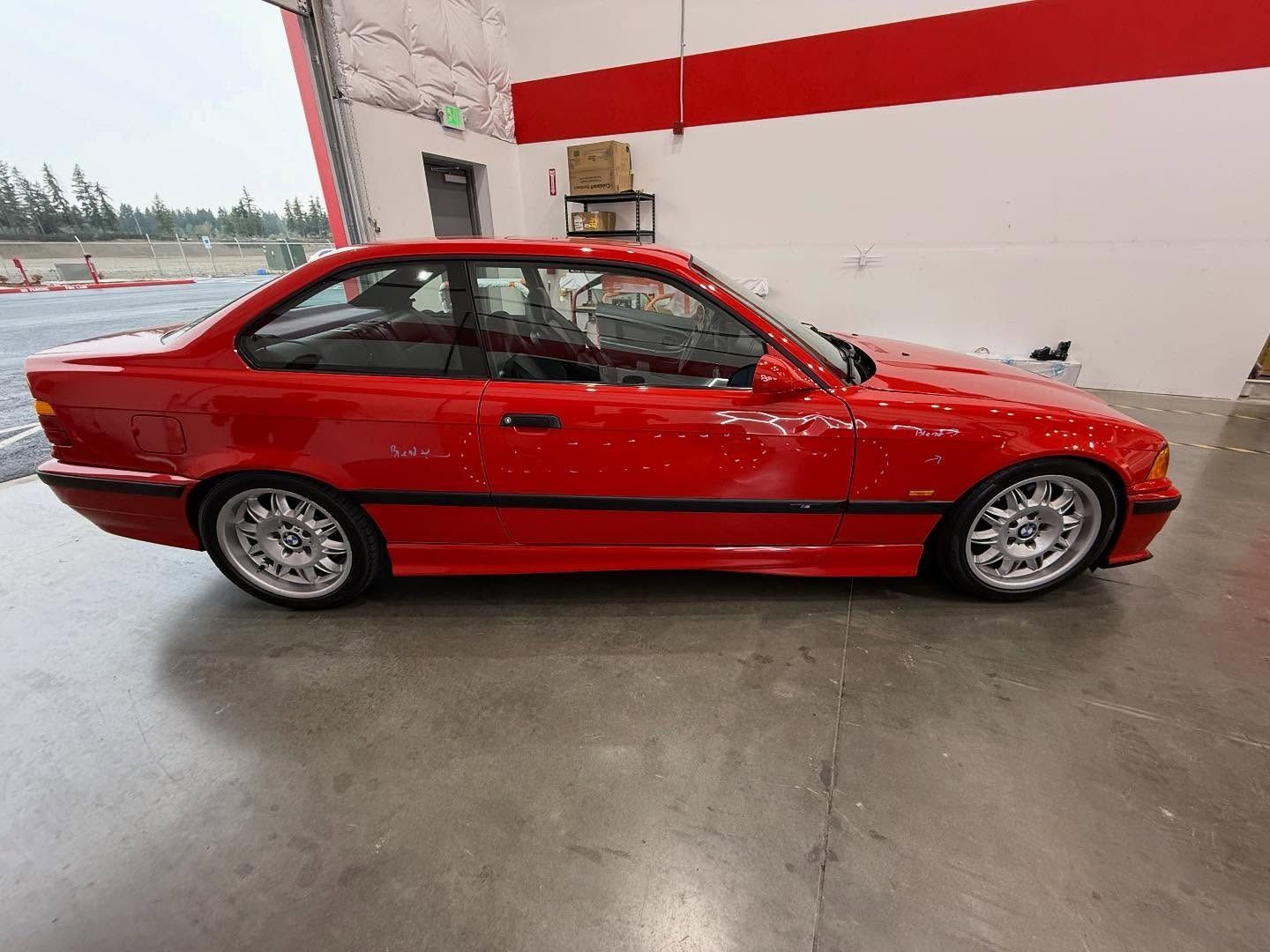 Red BMW E36 coupe parked inside a building with a red and white wall and grey flooring.