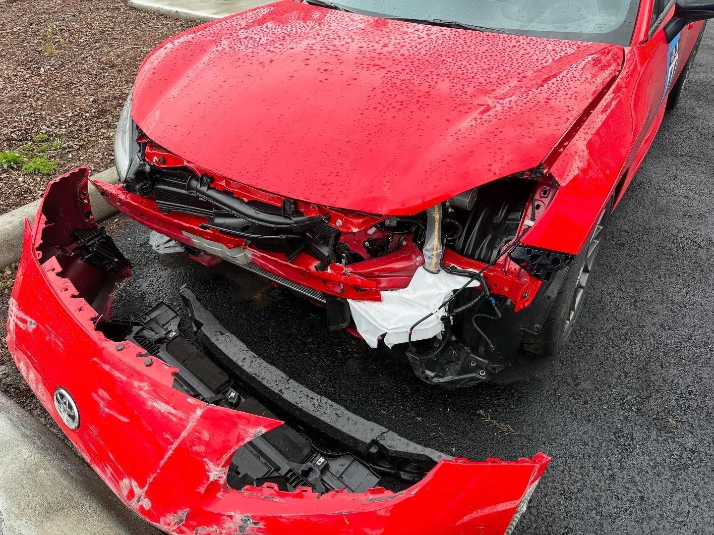 Damaged red car with a detached front bumper, showing significant front-end collision damage.