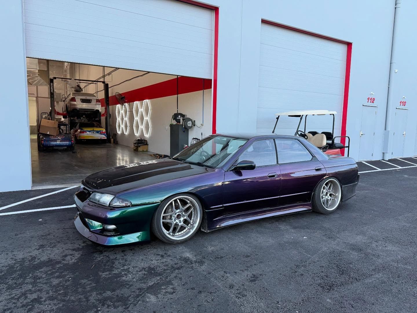 Nissan Skyline sedan with iridescent paint, parked outside a garage.