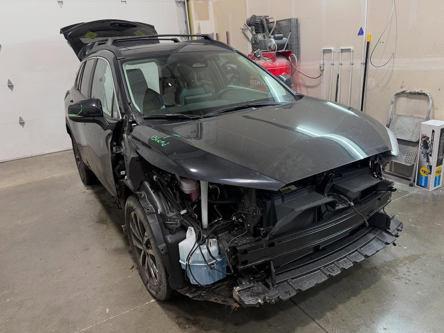 Black SUV with front-end damage; hood open, trunk open; in a garage.