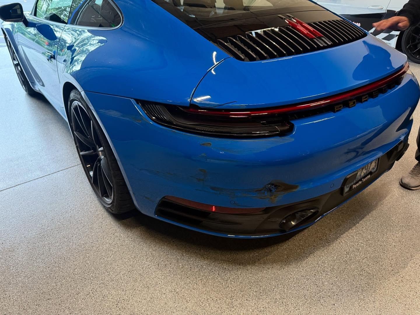 Blue Porsche sports car with rear damage; black wheels, dark taillights, black rear grill, and red stripe.