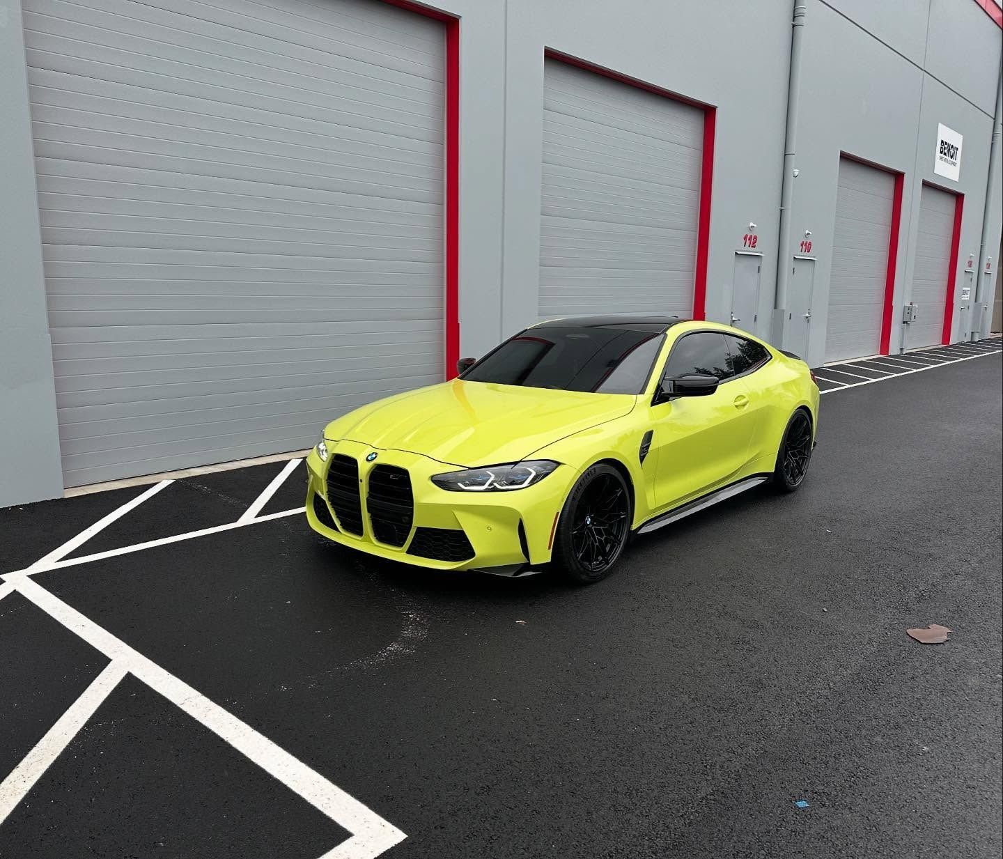 Yellow BMW M4 coupe parked on asphalt, black wheels, against gray building with red trim.
