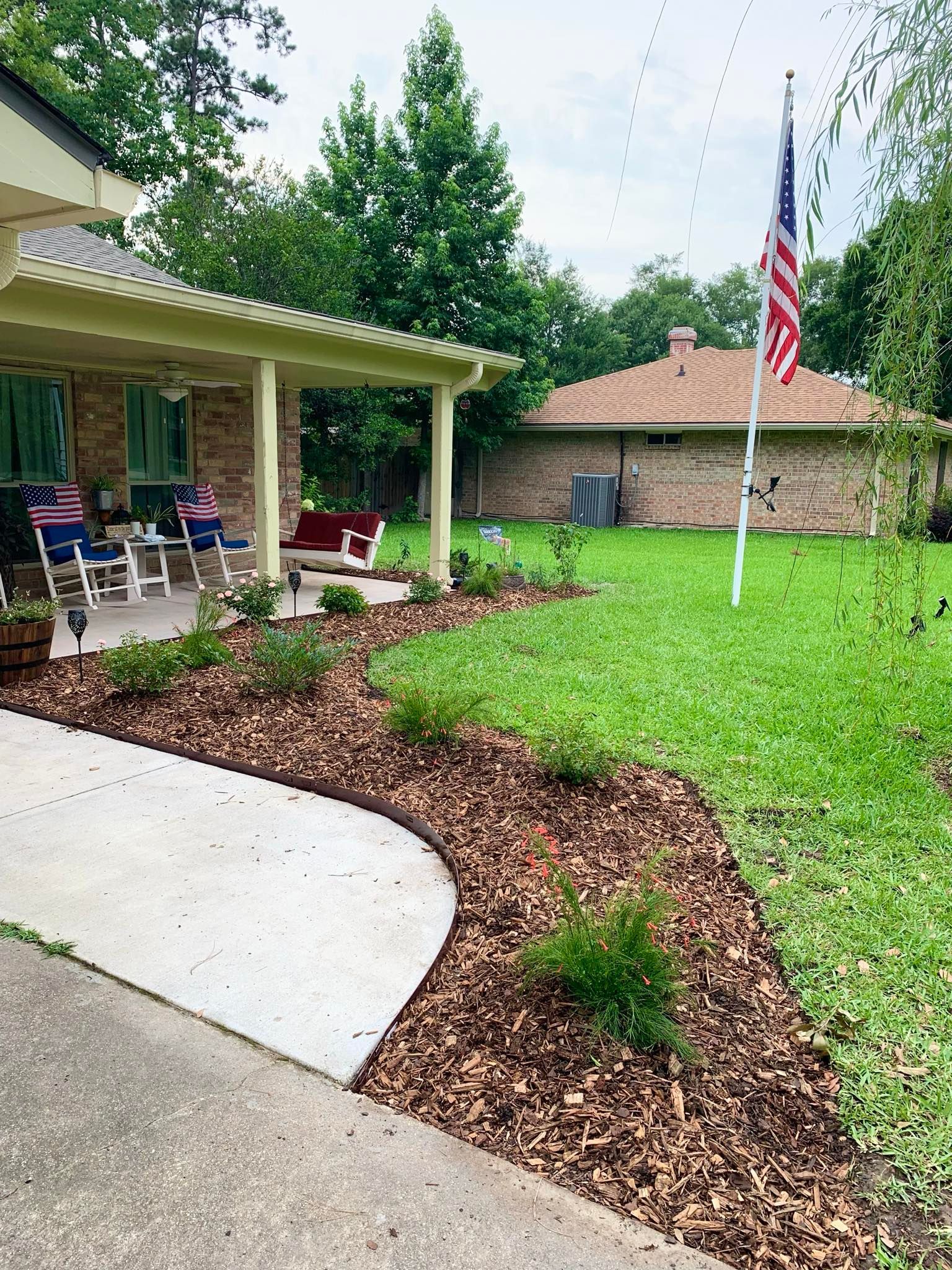 Covered front porch, curved concrete walkway, mulch beds, green lawn, and an American flag by a brick house.