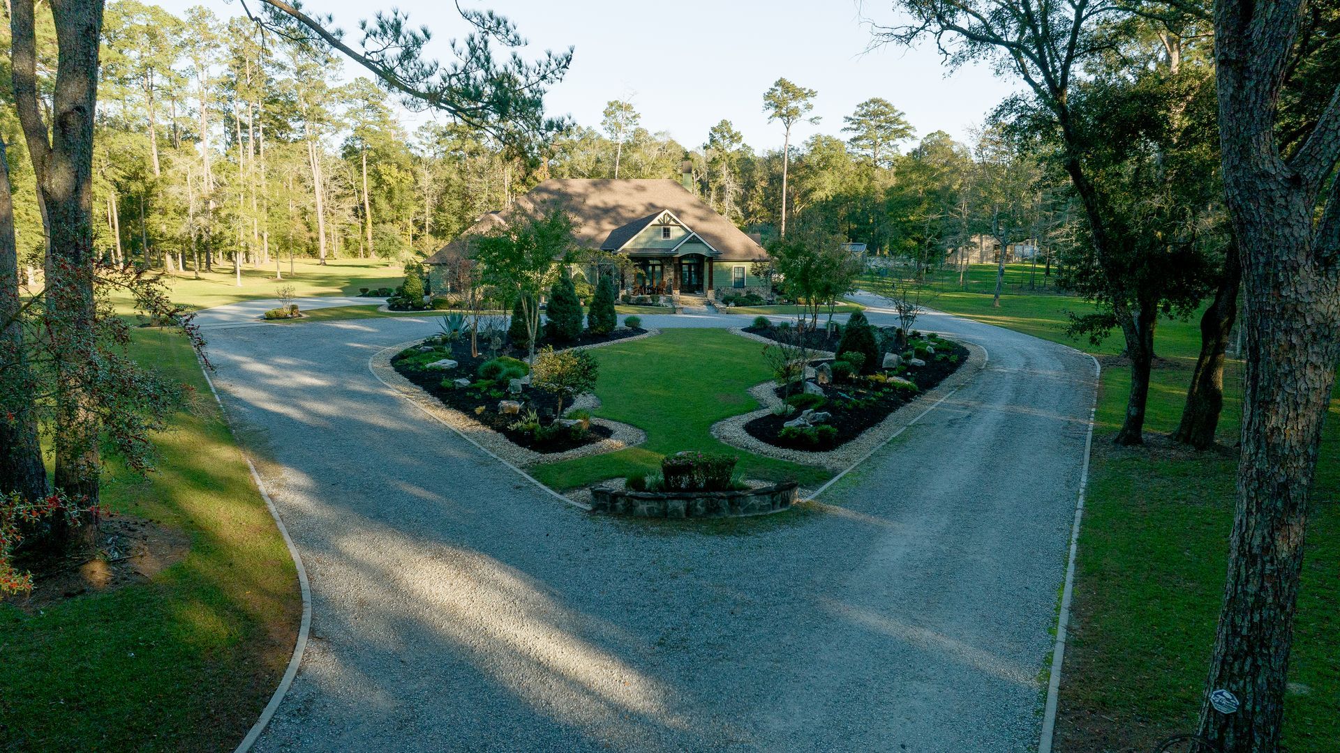 Curved gravel driveway around a landscaped garden leading to a house in a wooded setting