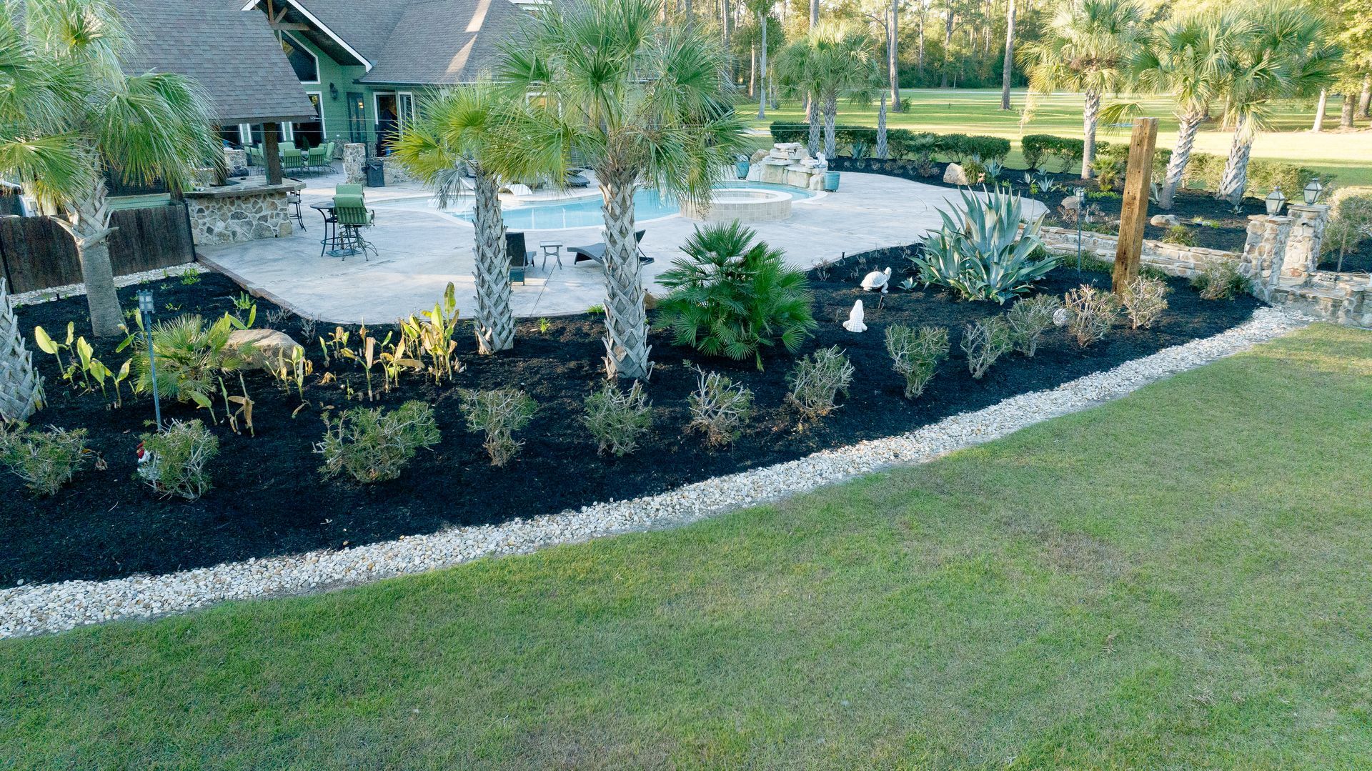Landscaped garden bed with shrubs, trees, and white stone edging beside a lawn and patio