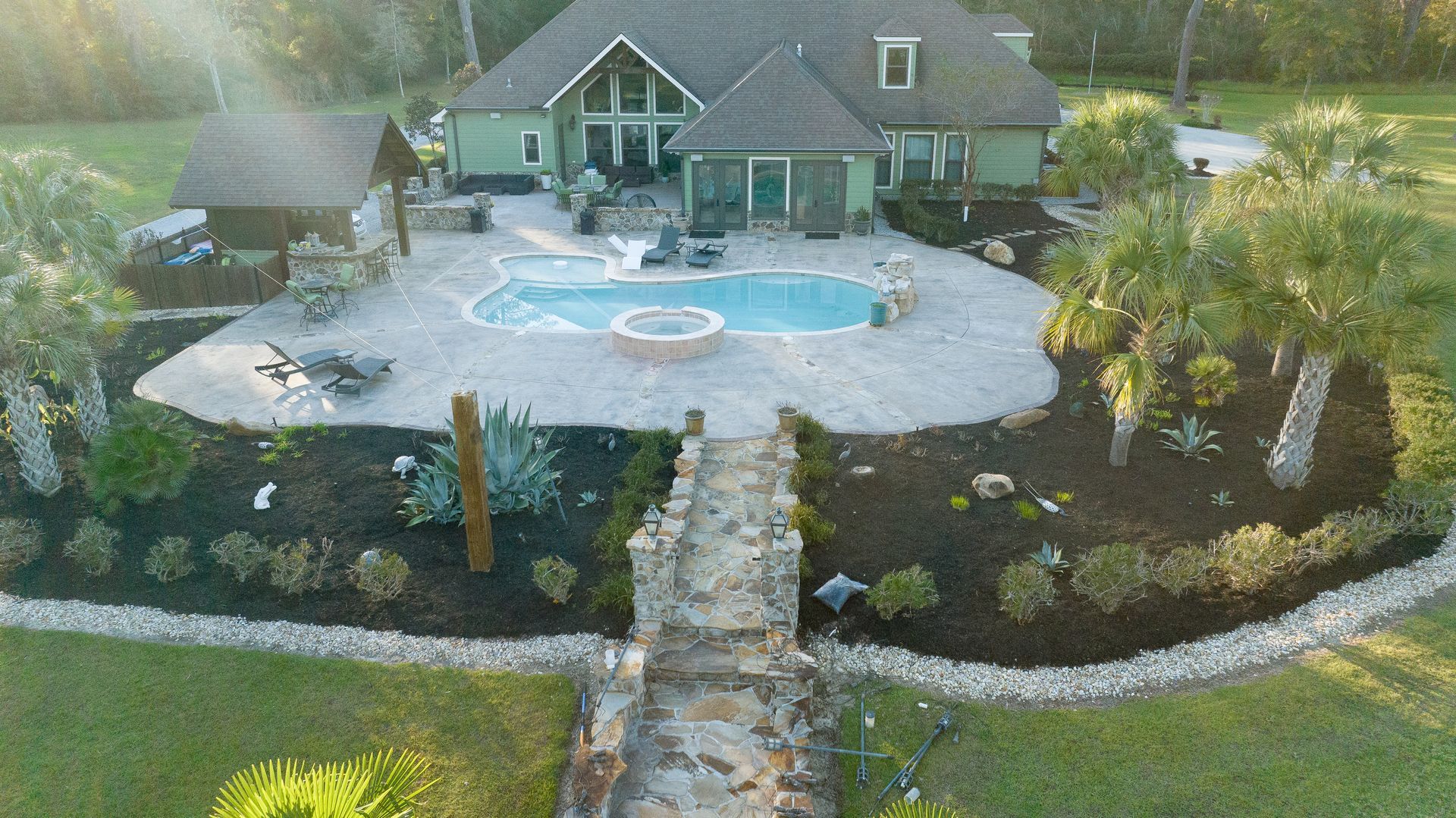 Aerial view of a house with a backyard pool, stone patio, and landscaped garden at sunset