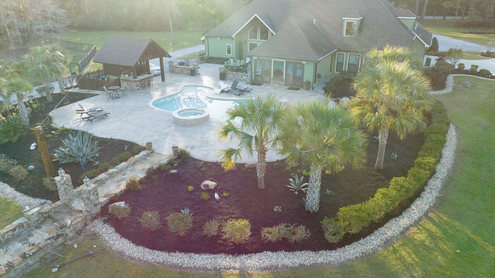 Aerial view of a landscaped backyard with a pool, stone patio, flower beds, and a large house.