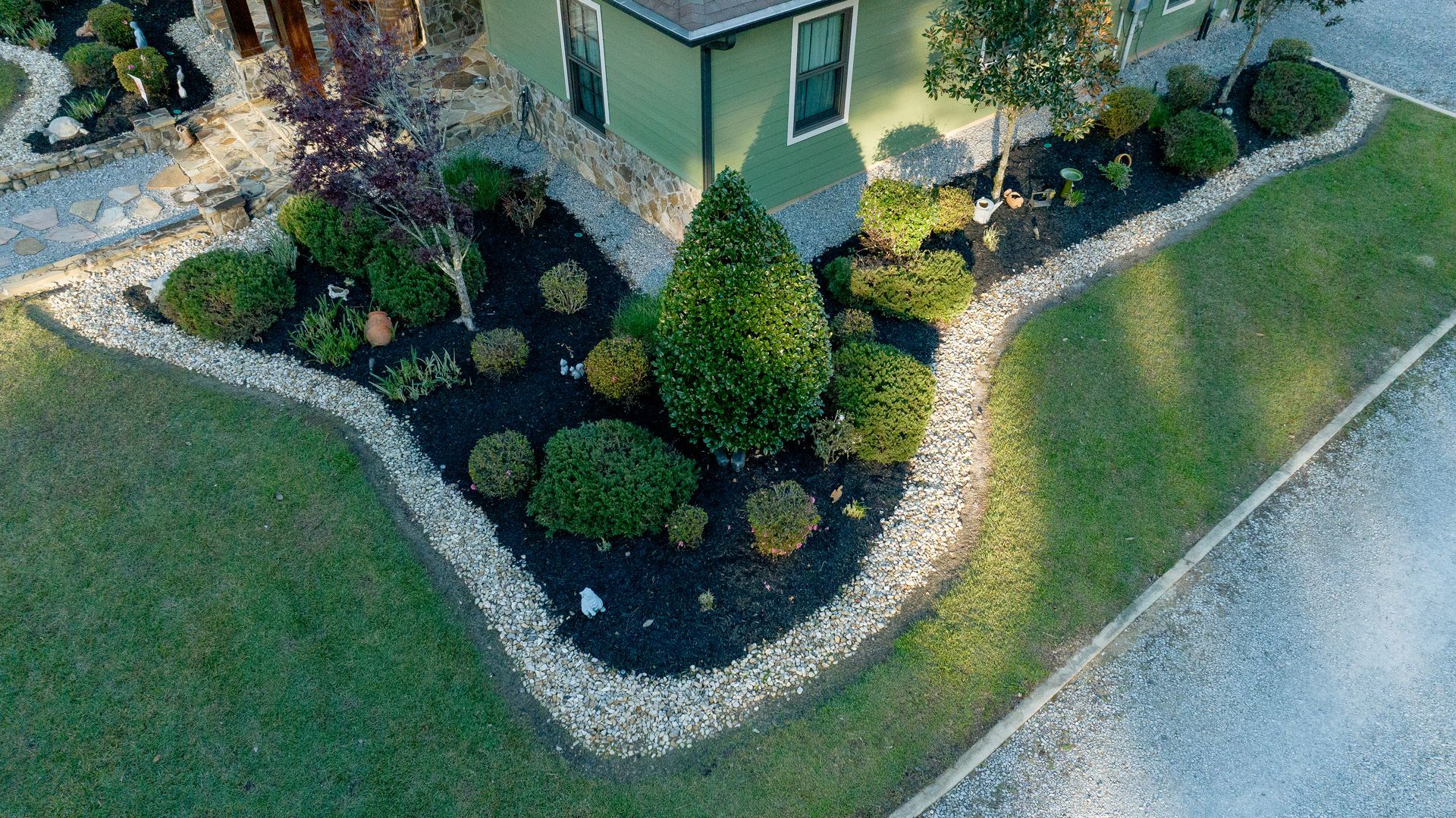 Aerial view of a house corner with landscaped shrubs, a stone border, and a gravel path beside grass.
