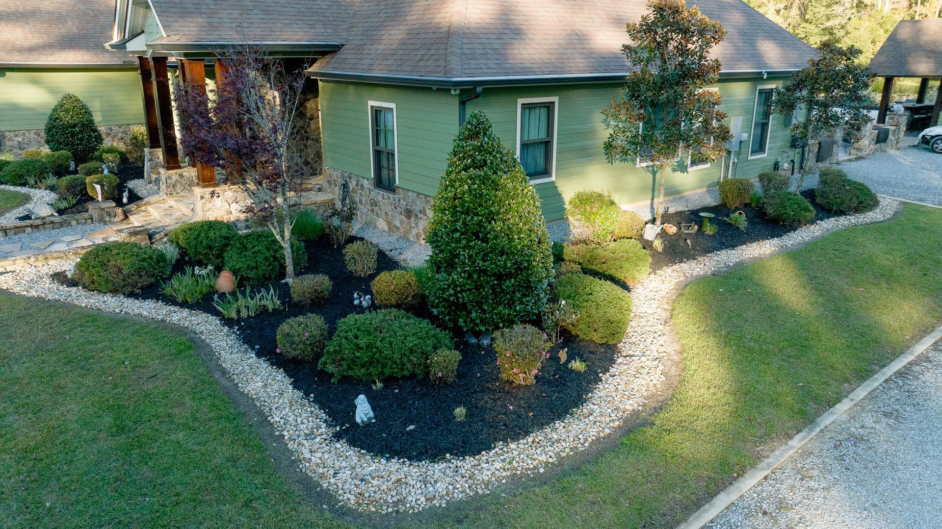Landscaped front yard with shrubs, mulch beds, and a green house with stone trim