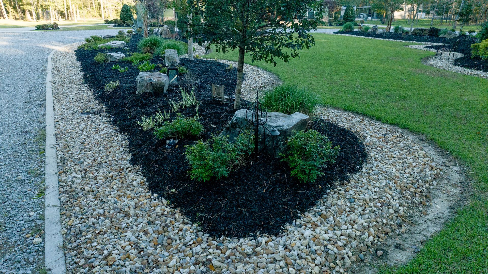 Landscaped garden bed with small shrubs and mulch, bordered by white gravel and a green lawn.