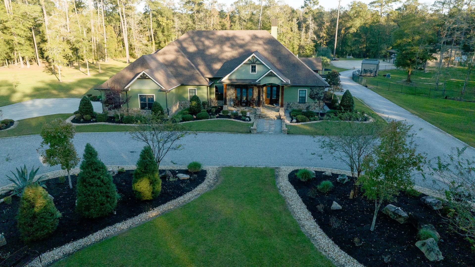 Stone house with a large front yard, curved gravel driveway, and landscaped flower beds surrounded by trees.
