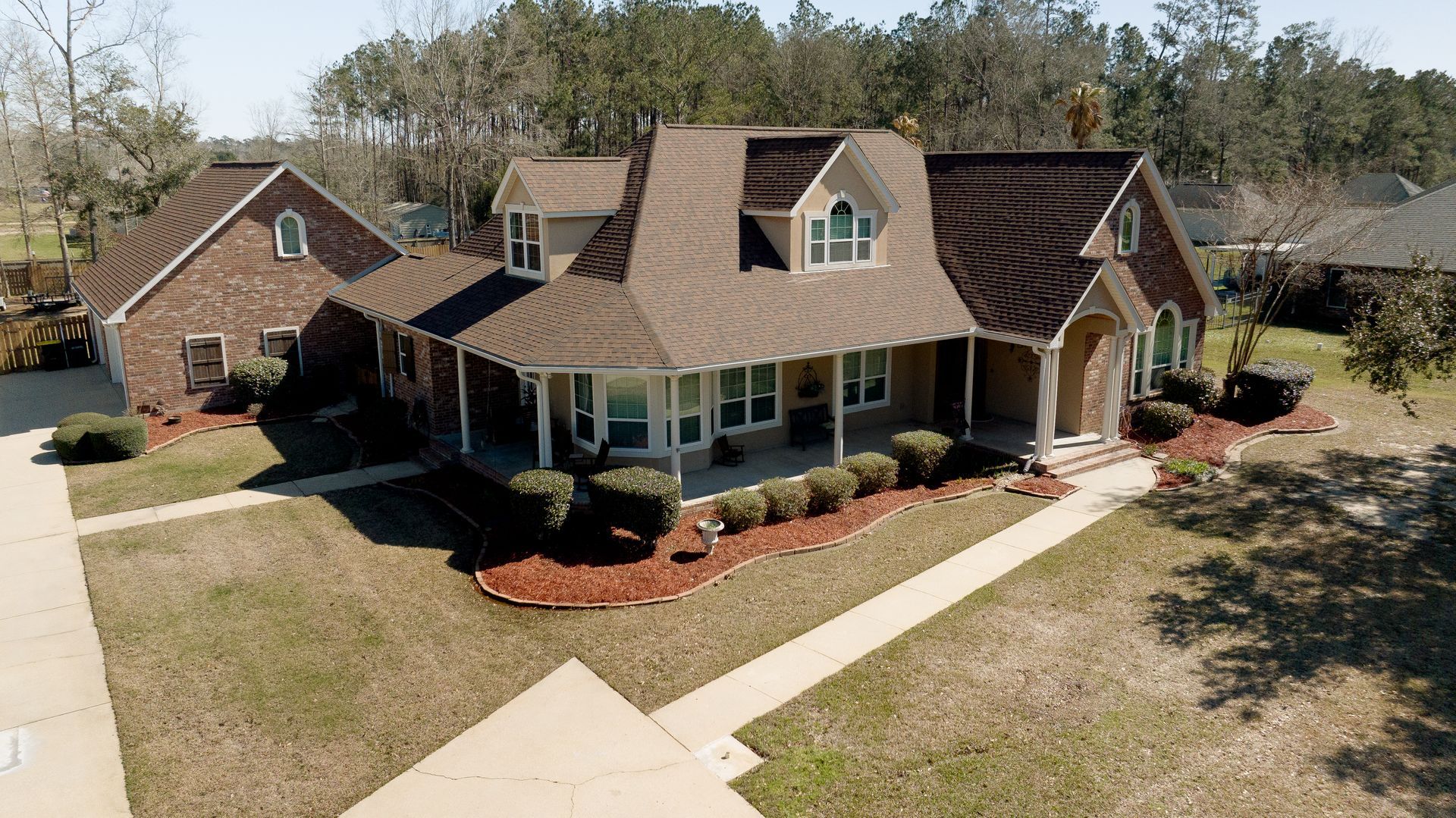 Aerial view of a suburban house with a brown roof, wraparound porch, and landscaped yard
