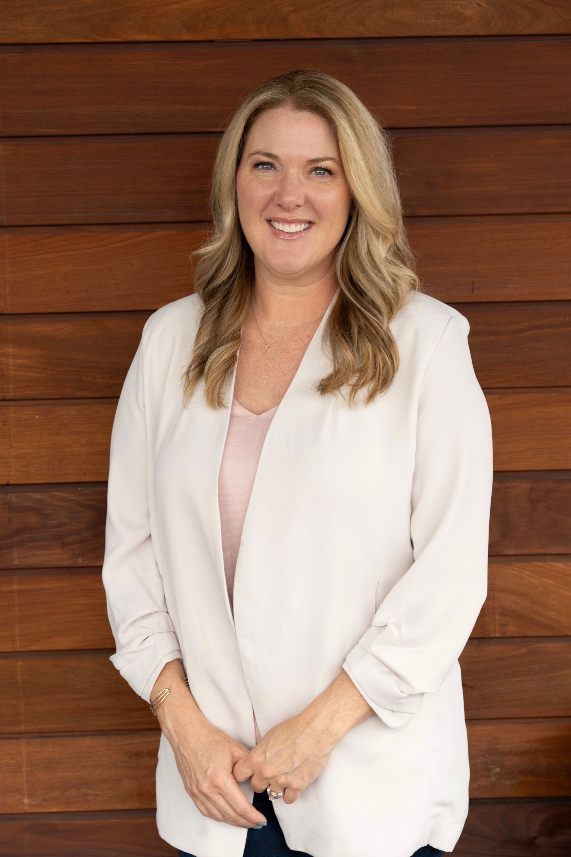 Woman smiles, poses against a wood wall, wearing a cream blazer and a pink top.