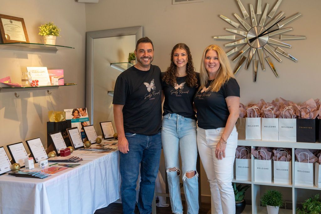 Three people smiling in a shop: a man, a young person, and a woman. They stand near a table and shelves.