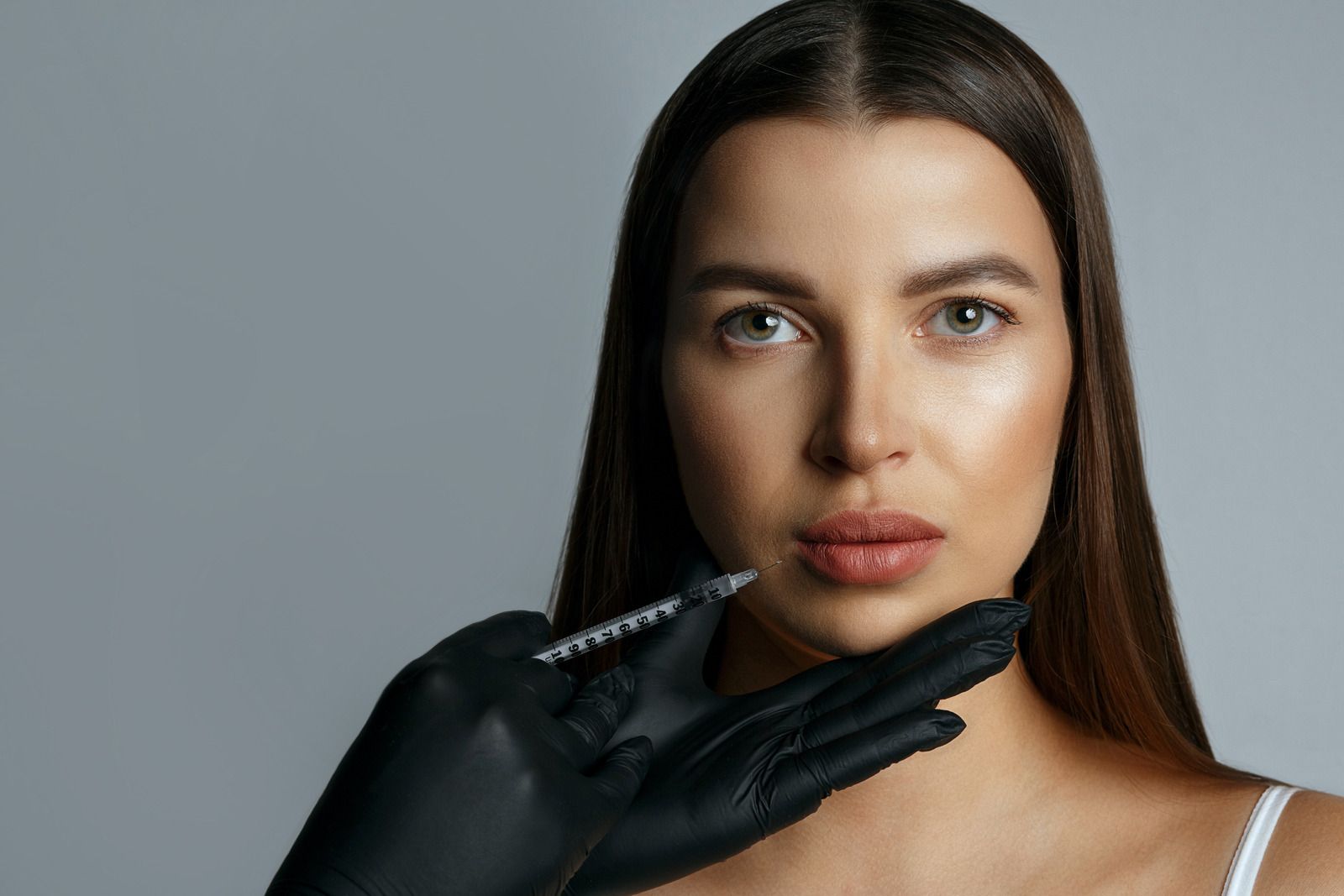Woman receiving lip filler injection. Black gloved hand holds syringe near lips. Gray background.