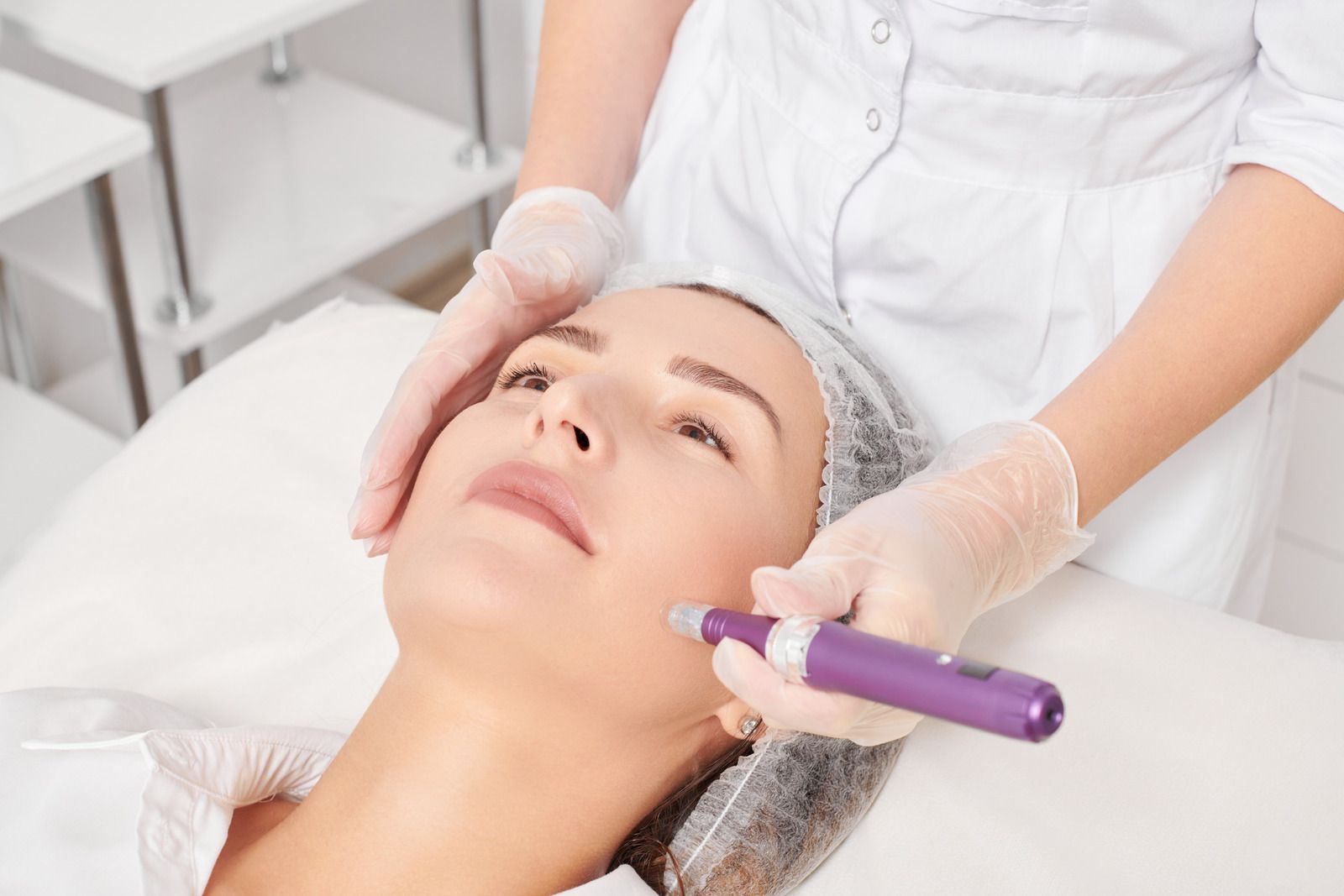 Woman receiving a microneedling treatment on her cheek, by a person wearing gloves in a clinic.