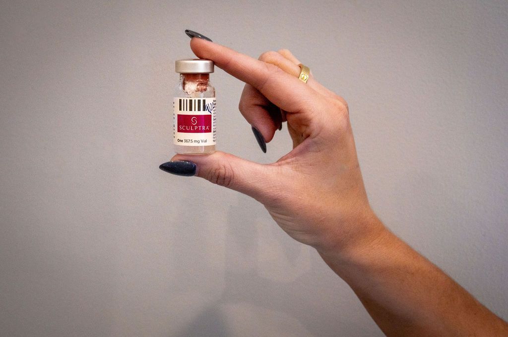 Hand holding a small vial with a red and white label, against a gray background.