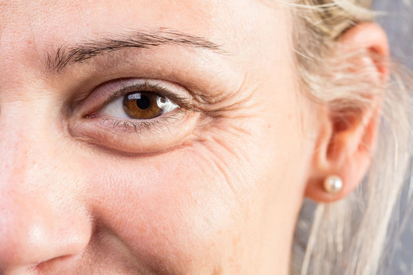 Close-up of a smiling eye and cheek with light makeup, blonde hair, and a pearl earring