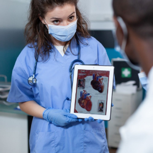 A healthcare worker in blue scrubs and a face mask holds a tablet displaying an anatomical illustration of a human heart.