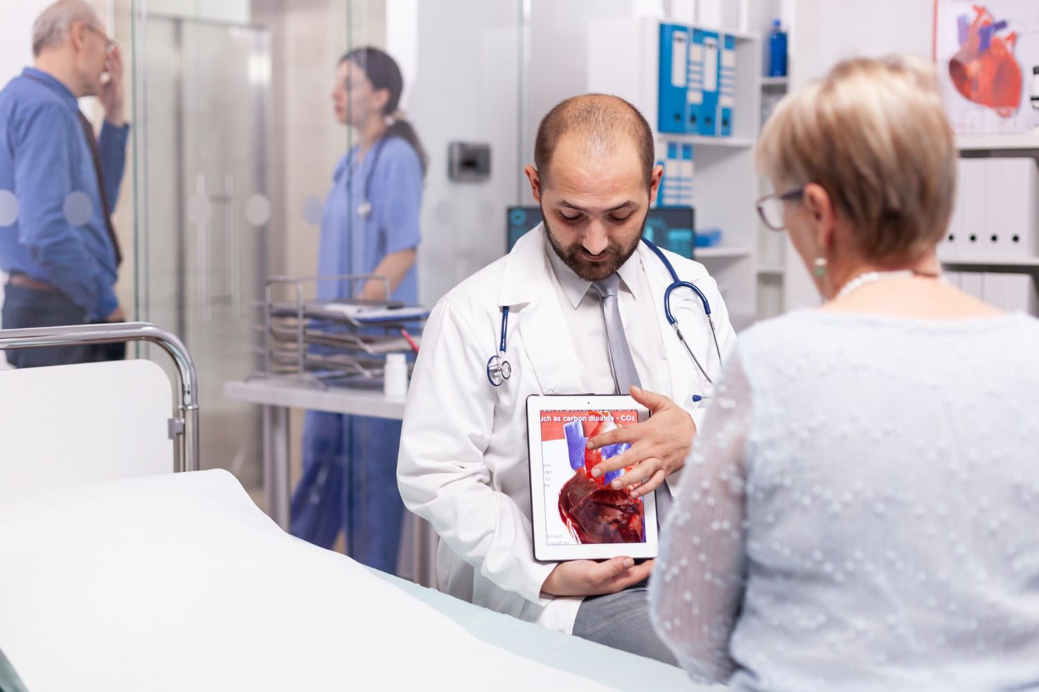 A doctor in a white coat shows a medical diagram of a heart on a tablet to a patient in a clinic office.