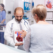 A doctor points to a heart model on a tablet while consulting with a patient in a medical office.