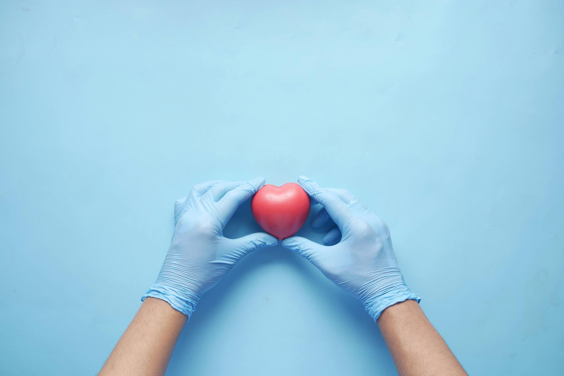 Two hands wearing light blue medical gloves hold a small, red, heart-shaped object against a light blue background.