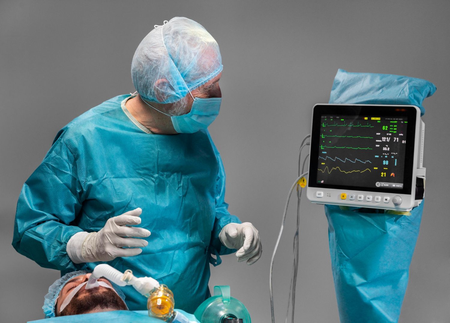 A masked medical professional in scrubs monitors a patient connected to a ventilator and a heart rate display.