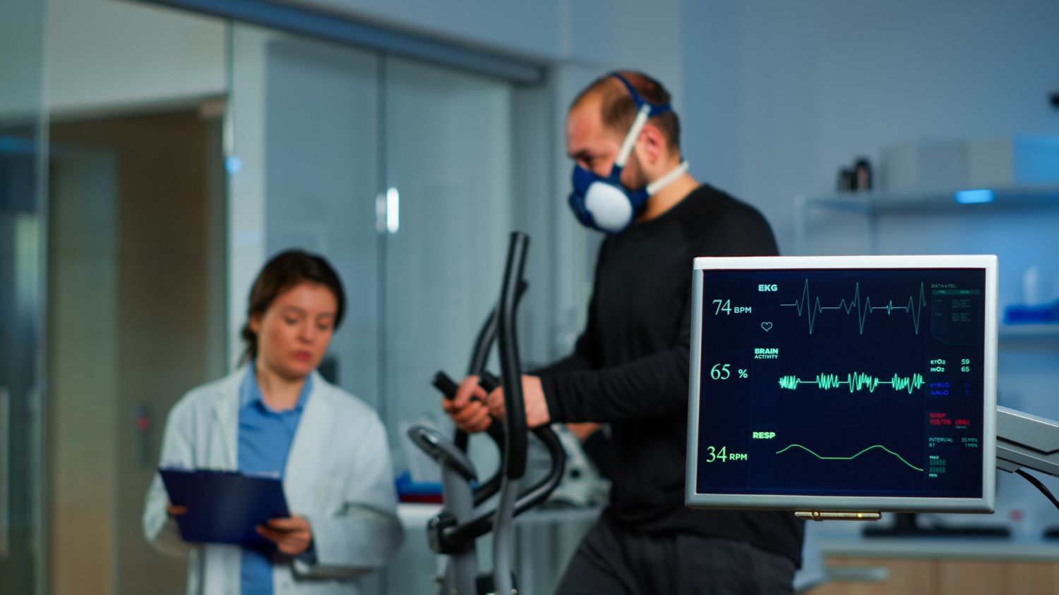 A person wears a mask while exercising on a machine in a lab as a professional monitors their vitals on a screen.