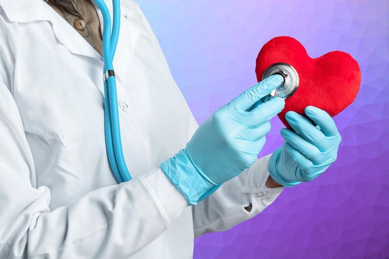 A healthcare professional in a white coat and blue gloves uses a stethoscope to examine a plush red heart.
