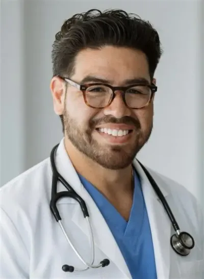 Doctor in a white coat with arms crossed, standing against a light gray background.