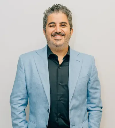 Smiling man in a light blue blazer and dark shirt against a plain light background