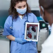 A healthcare worker in blue scrubs and a face mask holds a tablet displaying an anatomical illustration of a human heart.