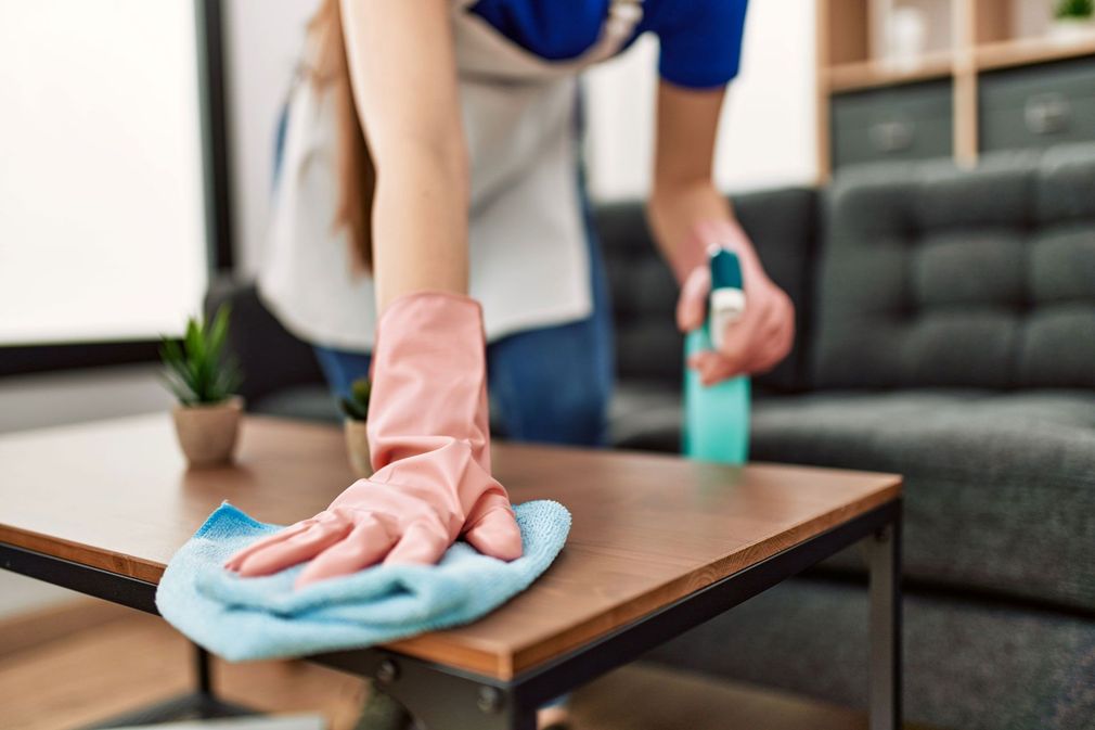 A person wearing pink rubber gloves wipes a wooden coffee table with a blue cloth while holding a spray bottle.